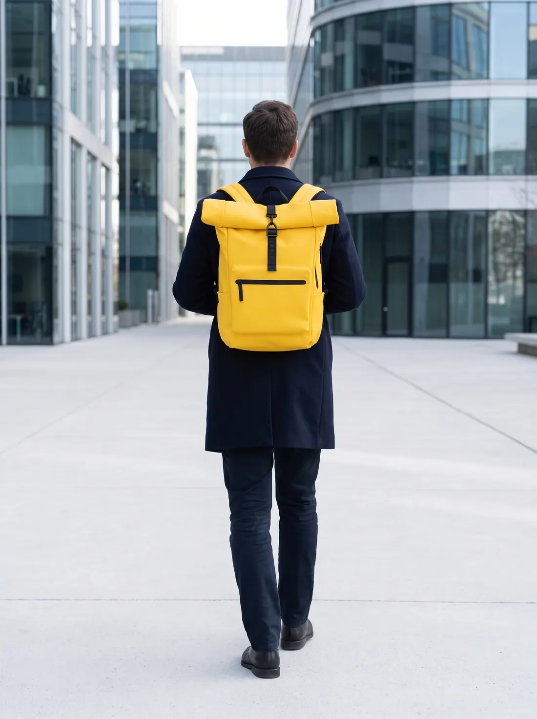 Eine Person mit dem Rücken zur Kamera überquert einen Büroplatz mit einem gelben Pilatus Urban Rucksack
